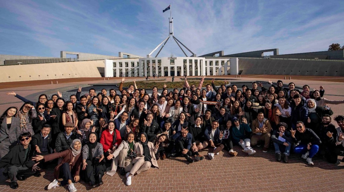 The photo captures a large group of participants seated on the ground in front of Australia's Parliament House in Canberra in the year 2023. The participants are smiling, showcasing a sense of camaraderie and engagement during their gathering at this iconic location
