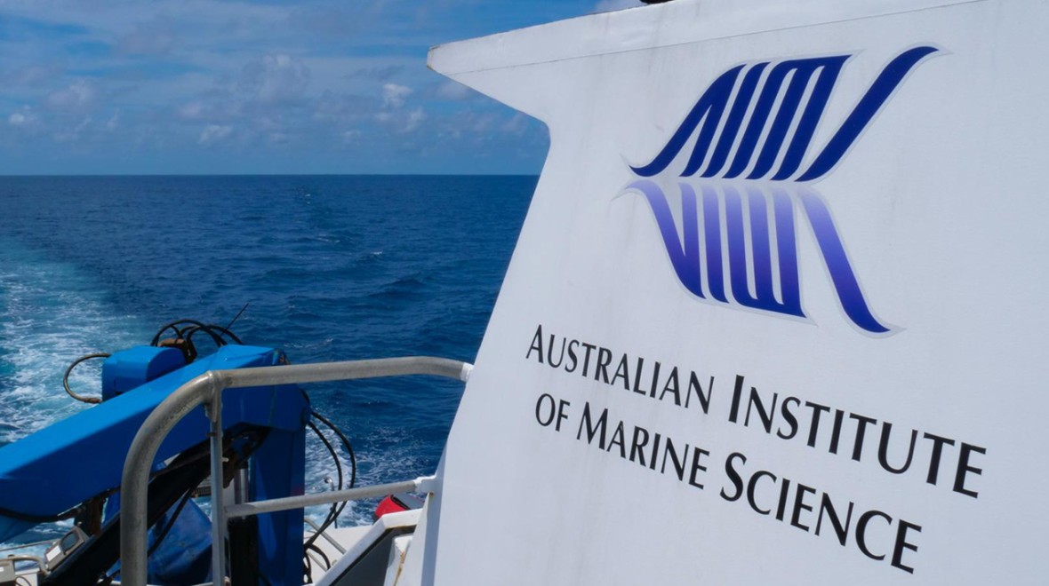 The image captures the back of a ship featuring the inscription 'Australian Institute of Marine Science.' The accompanying logo displays the initials of each word, and their reflection beneath forms a creative representation resembling a fish shape..