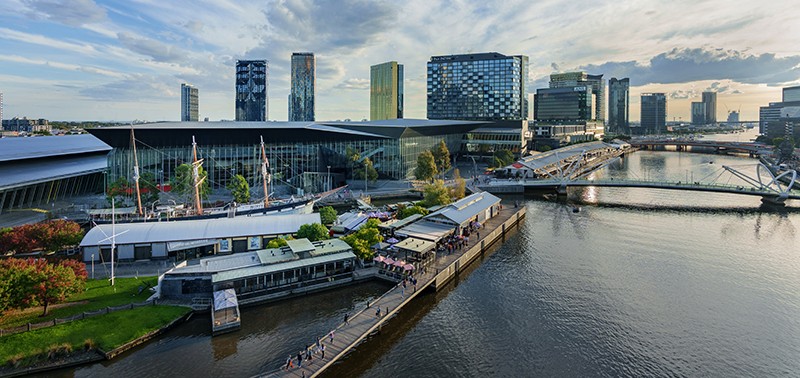The photo captures the Melbourne Convention and Exhibition Centre, featuring a scenic view. On the right-hand side, a river with two bridges is visible, while at the bottom, people are walking towards what appears to be the waterfront. Tall buildings in the background stand behind the exhibition centre, creating a dynamic cityscape. This image encapsulates the urban landscape and vibrant atmosphere around the Melbourne Convention and Exhibition Centre.