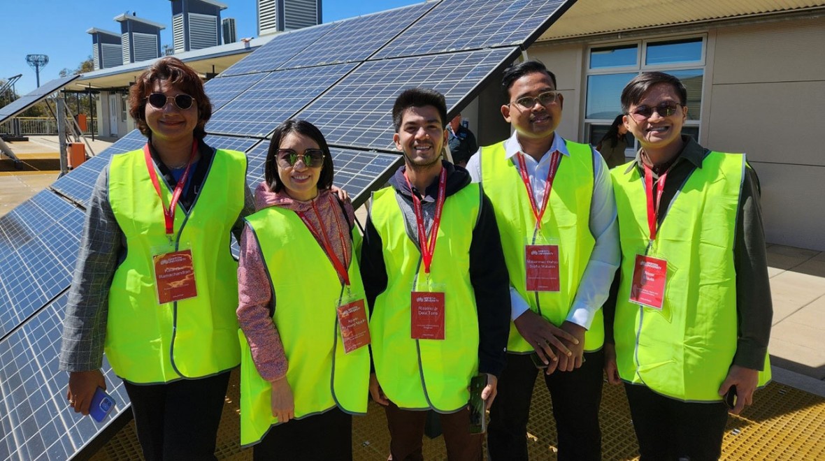 This photo depicts five individuals wearing safety vests and name tags, standing in front of solar panels on the roof of a building during a 2023 site visit to Canberra Institute of Technology. The participants are engaged in learning about Australia's renewable energy technology, highlighting their interest and involvement in sustainable practices.