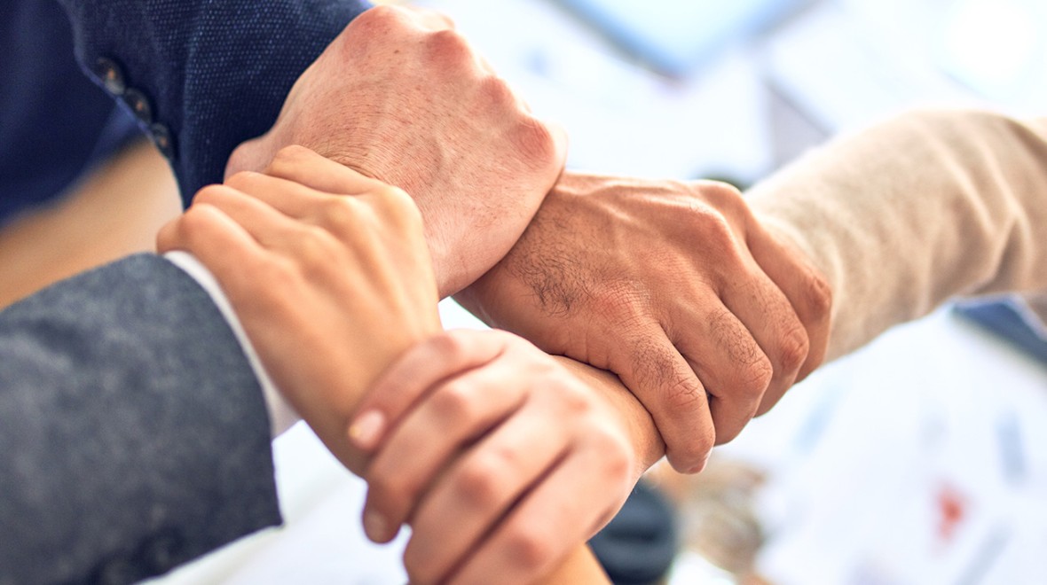 The image depicts four hands gripping together, forming an almost square shape. The hands belong to individuals of different ages and genders. The background is intentionally blurred, adding emphasis to the diverse unity symbolised by the hands coming together. This visual conveys a message of inclusivity and collaboration among individuals of varying ages and genders.