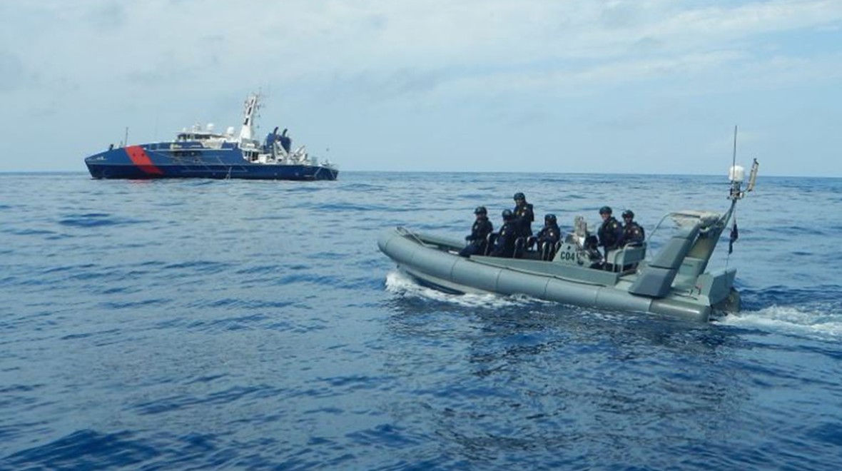 This image captures the dynamic scene of Border Force operations at sea, showcasing both the larger vessel and the speed boat in action. In the background, an Australian Border Force vessel is prominently displayed on the water. In the foreground, a smaller speed boat, also from the Australian Border Force, is heading towards the larger vessel. The speed boat carries six individuals in uniform, part of the Border Force team. 