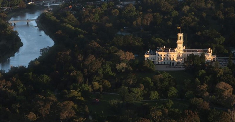 Aerial view of Government House Victoria white building constructed from 1872-1876. The central part includes a tall tower with a flagpole. Surrounding feature lush green trees, and to the left, a winding river with a bridge.